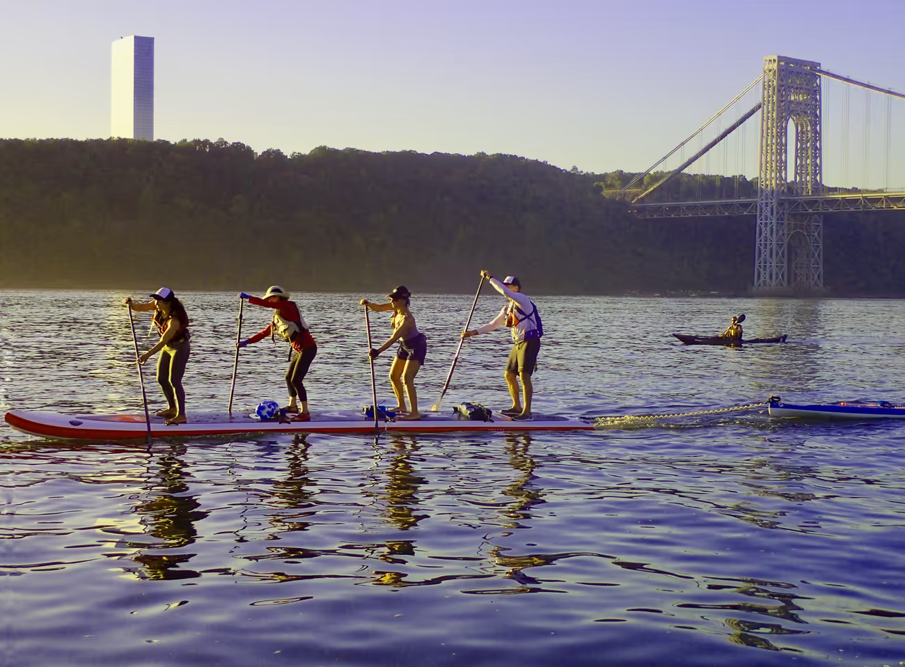 An inflatable four-person Red Paddle Co Dragon board paddling toward the George Washington Bridge at sunset.