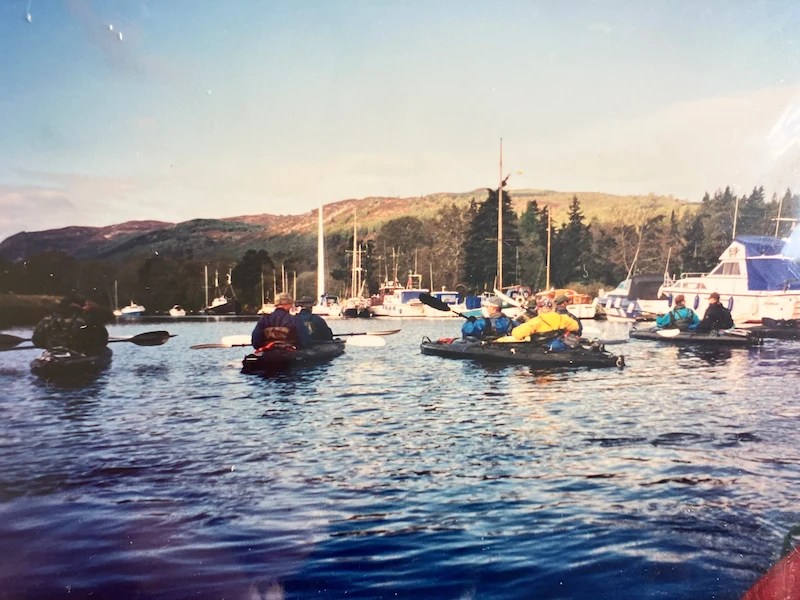 People kayaking in a calm lake with boats and trees in the background.