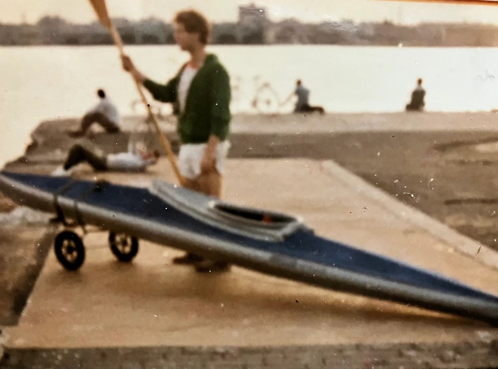 Person standing with a kayak on a dock near a river.