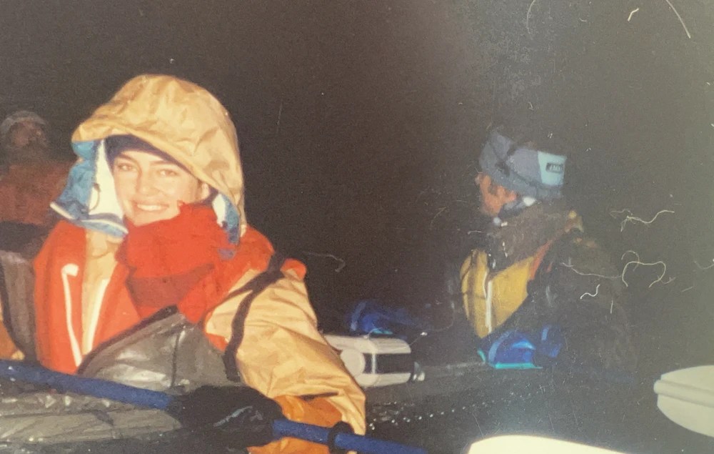 Two people in rain gear on a boat at night, one smiling at the camera.
