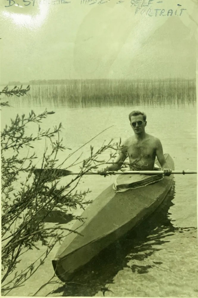 Shirtless man paddling a kayak on a lake near leafy branches.