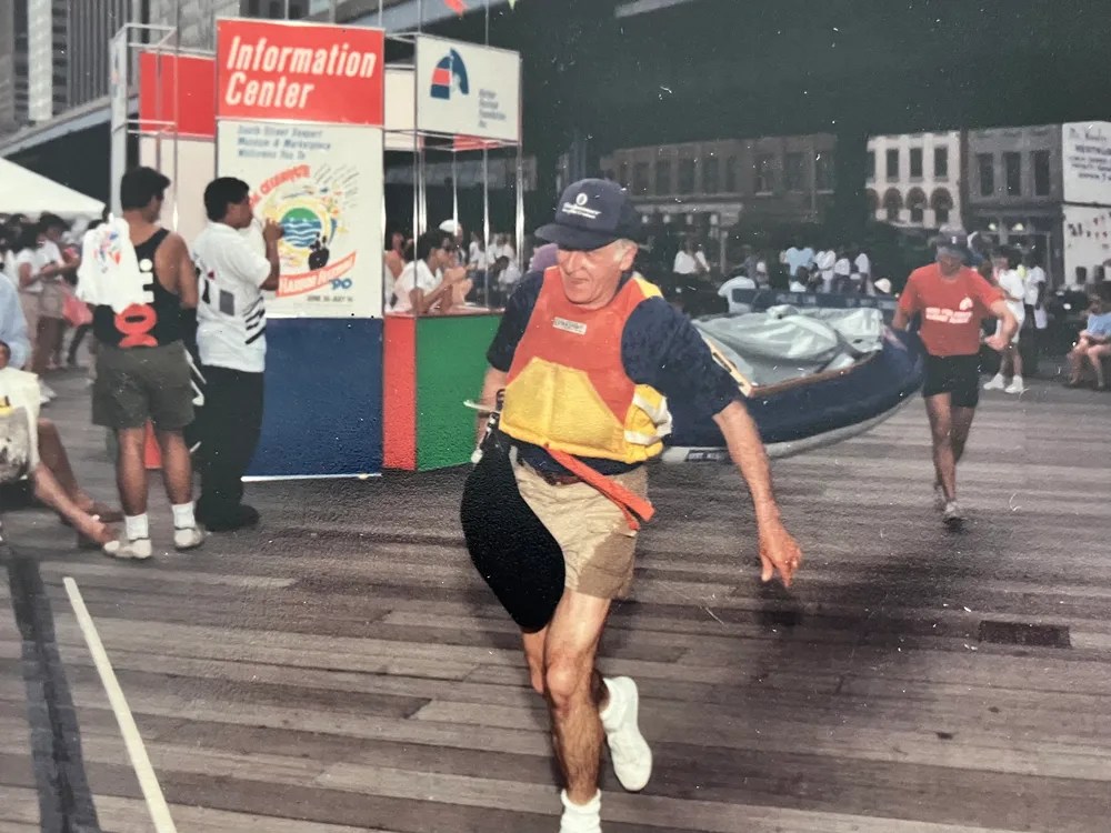 Man running on boardwalk near information center, wearing a cap and life vest, with people and tents around.