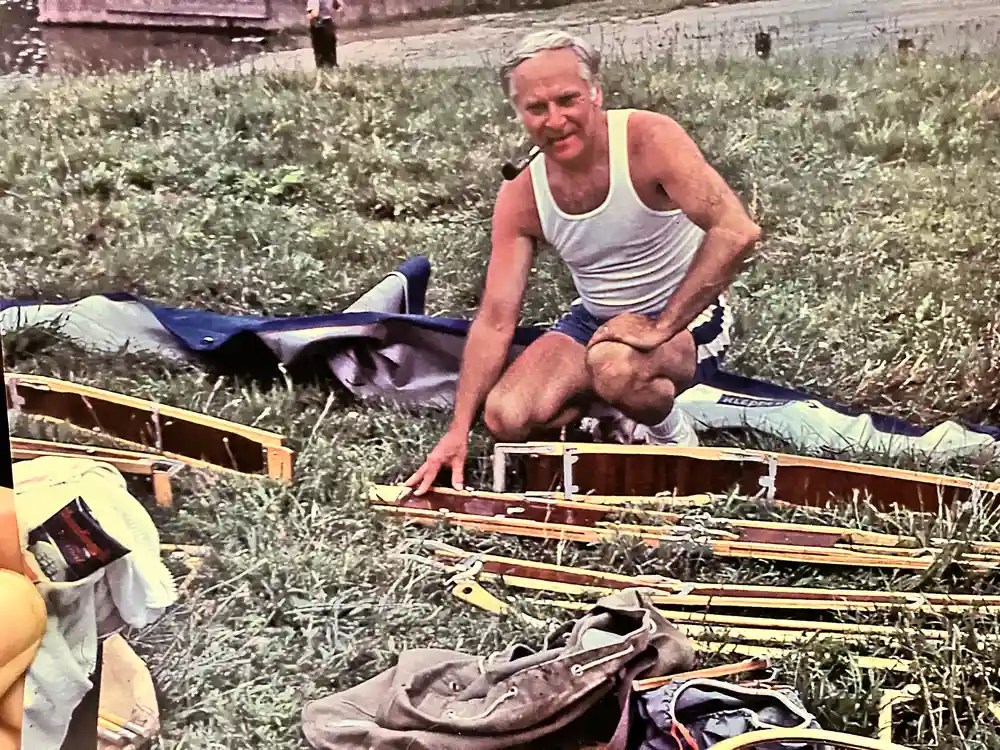 Man assembling a wooden structure, sitting on grass, wearing a tank top and shorts, smoking a pipe.