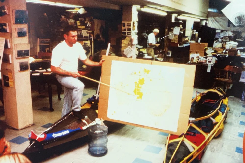 Man presenting a map in a cluttered room with a kayak nearby.