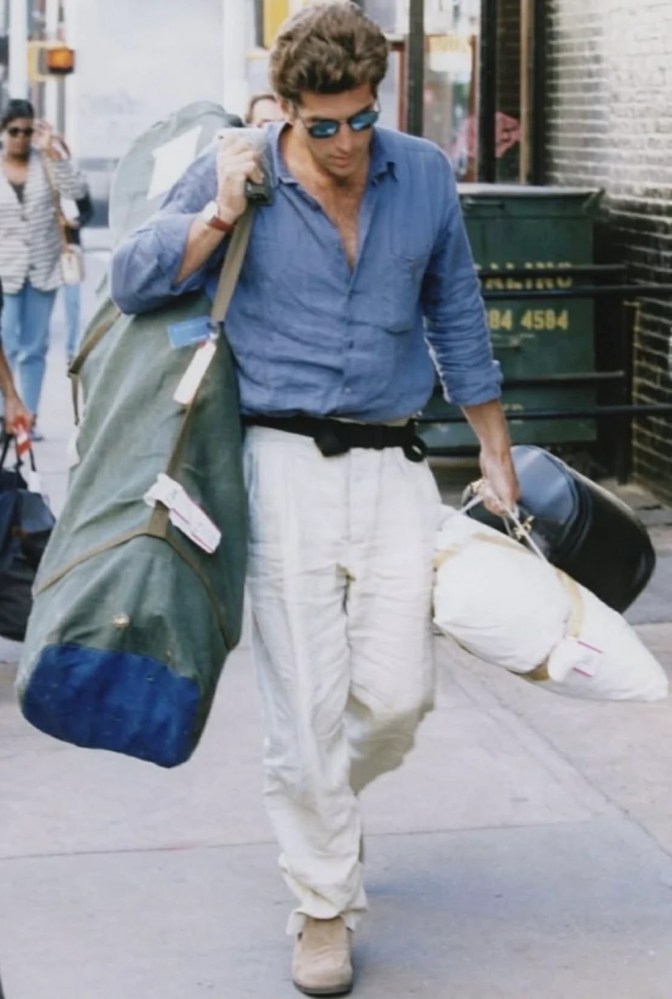 Person in blue shirt, white pants carrying large bags, walking on city sidewalk.
