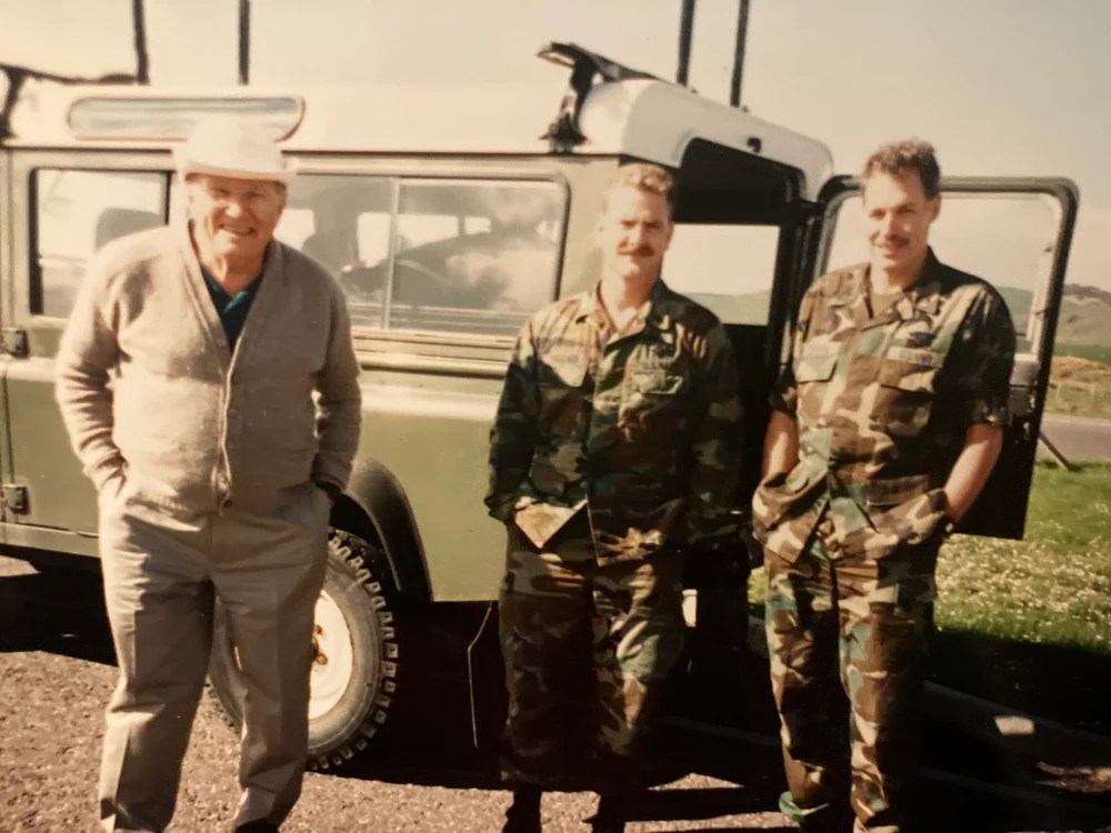 Three men in front of a vehicle, two in camo, one in a sweater.