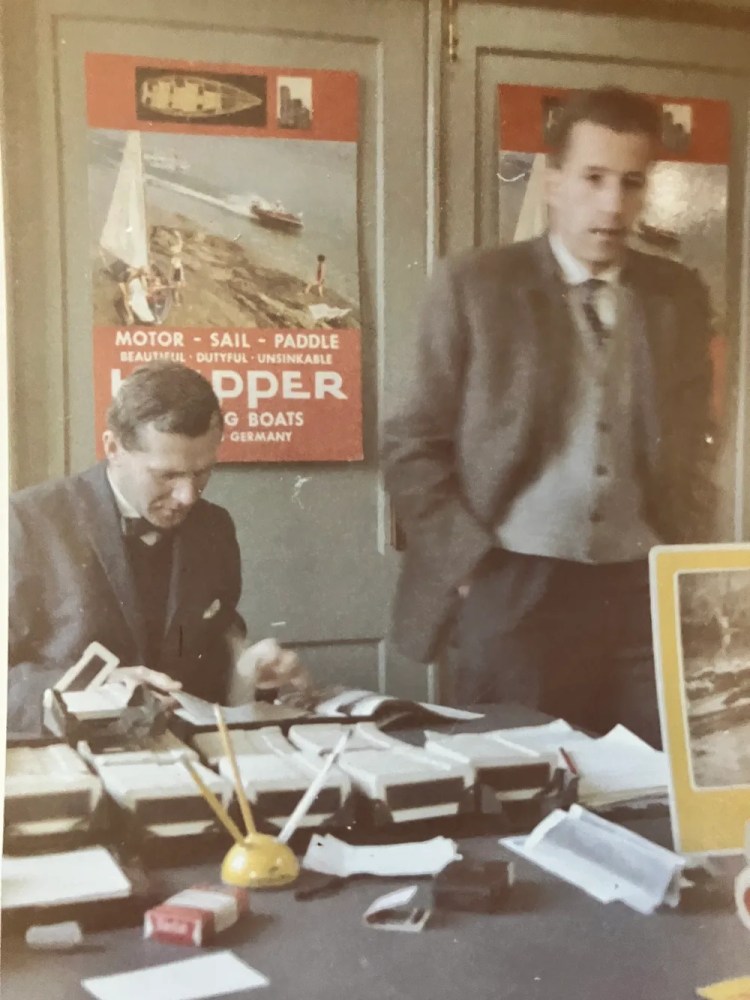 Two men in suits working at a cluttered desk with boating posters on the wall.
