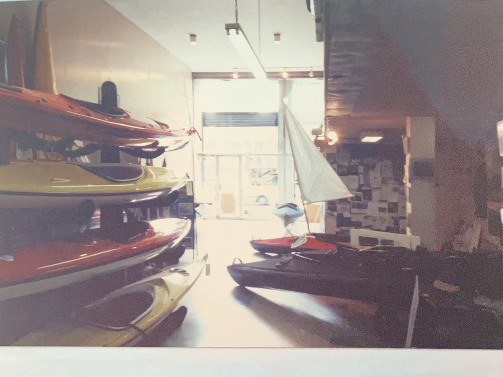 Indoor display of kayaks on racks and a small sailboat with a white sail.