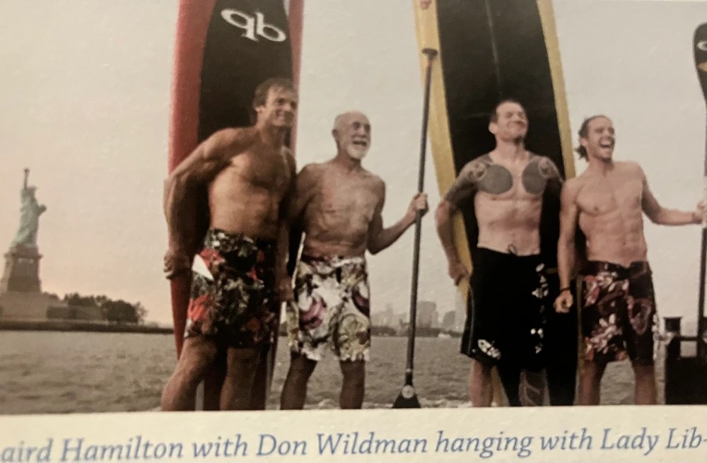 Four men in swimwear holding surfboards near the Statue of Liberty.
