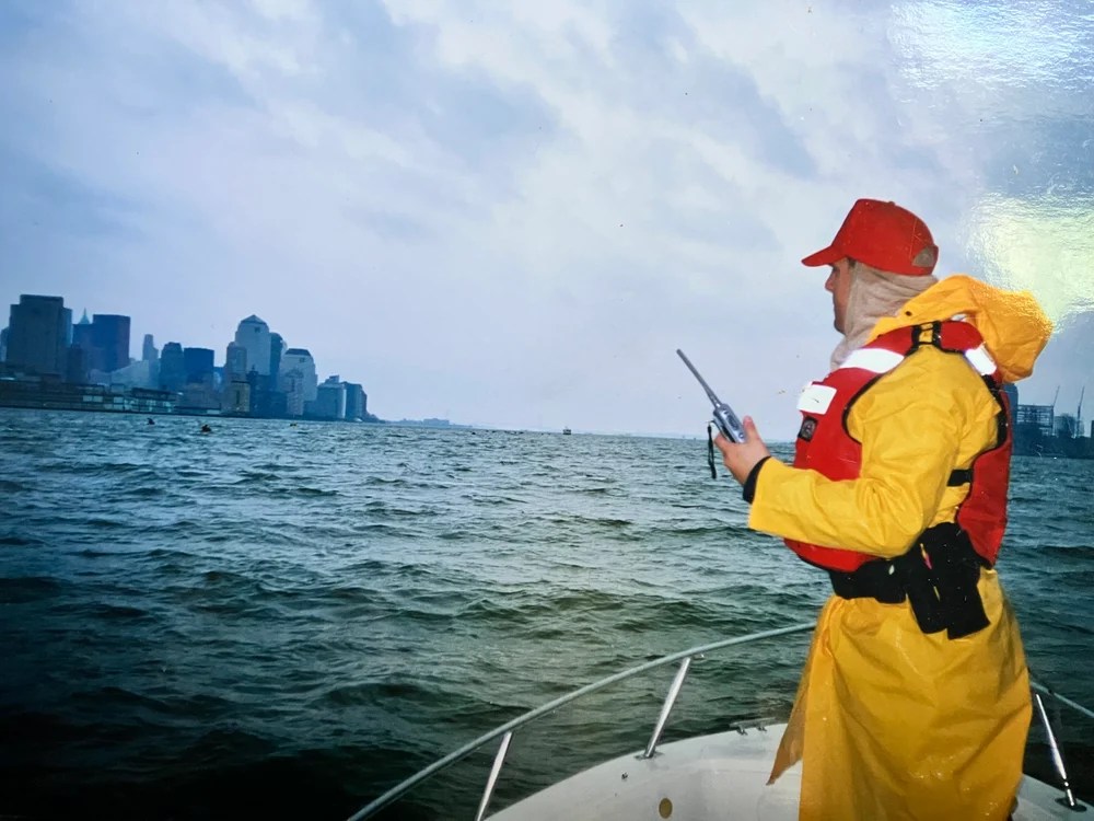 Person in yellow raincoat and red hat on a boat with city skyline in the distance.