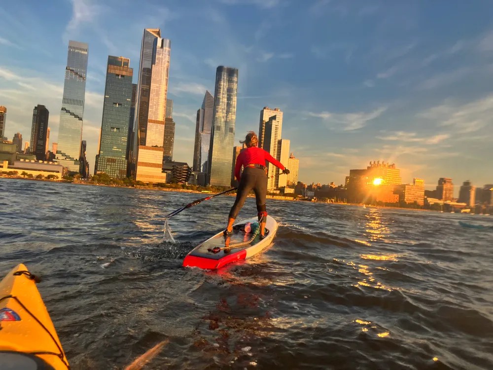 Person paddle boarding on river with city skyline and sunset.