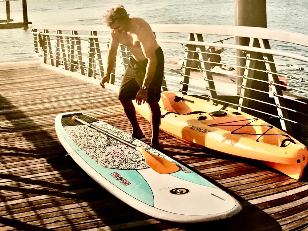 Man on pier with paddleboard and kayak in sunlight.