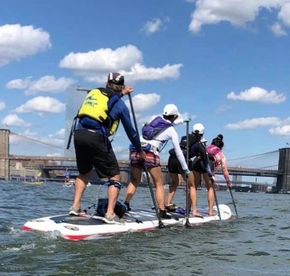 Four people paddle boarding on a large board in a river near a bridge.