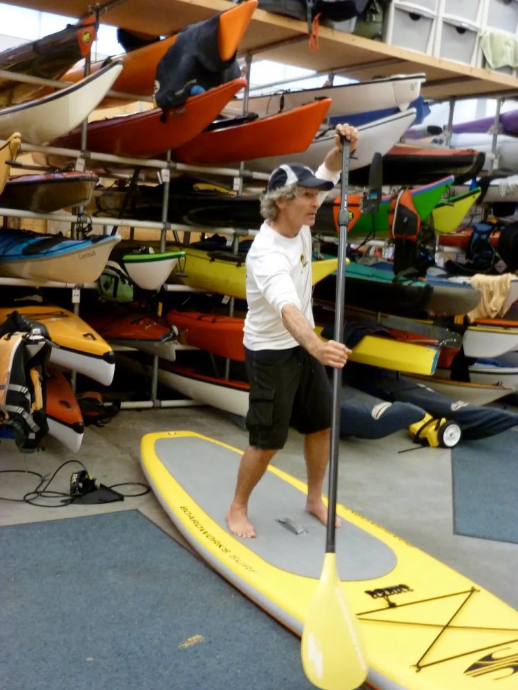 Person standing on a paddleboard indoors, surrounded by boats on racks.