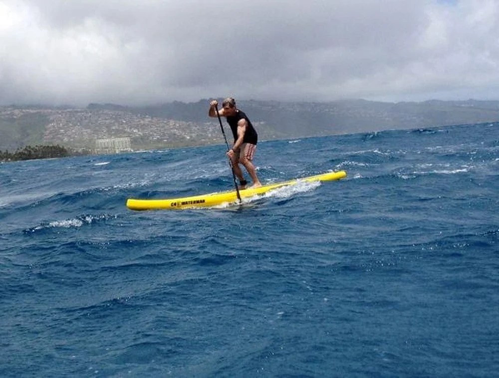 Person paddleboarding on rough ocean waters under cloudy skies near a hilly coastline.