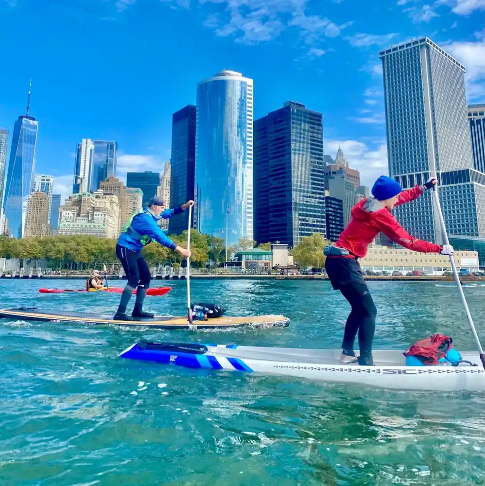 Two people paddleboarding on water with a city skyline in the background.