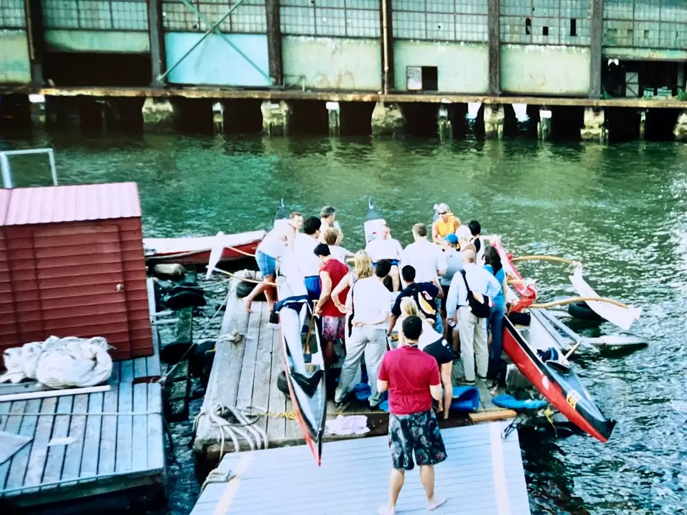 People gathered on a dock by the water with boats visible.