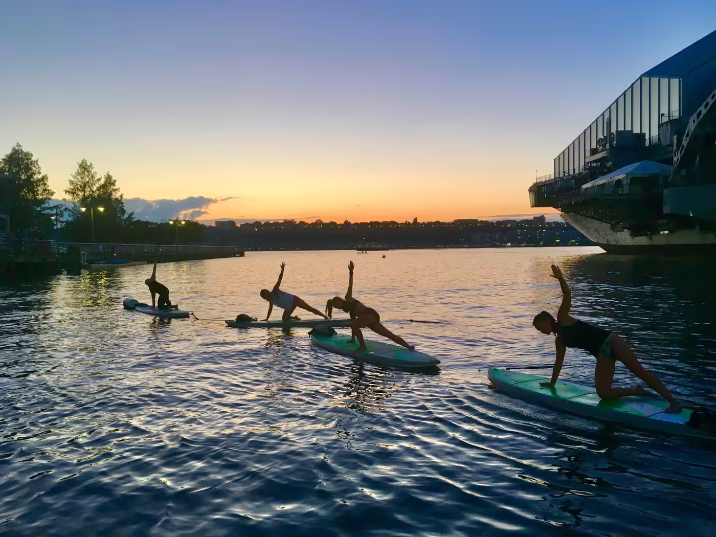 Four women doing a supported side plank on paddleboards during sunset at the Hudson River Park's Pier 84 Boathouse.