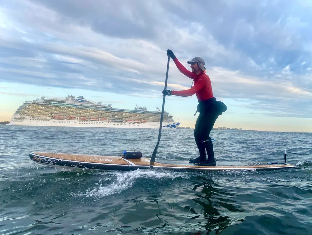 Person paddleboarding with a cruise ship in the background on a cloudy day.