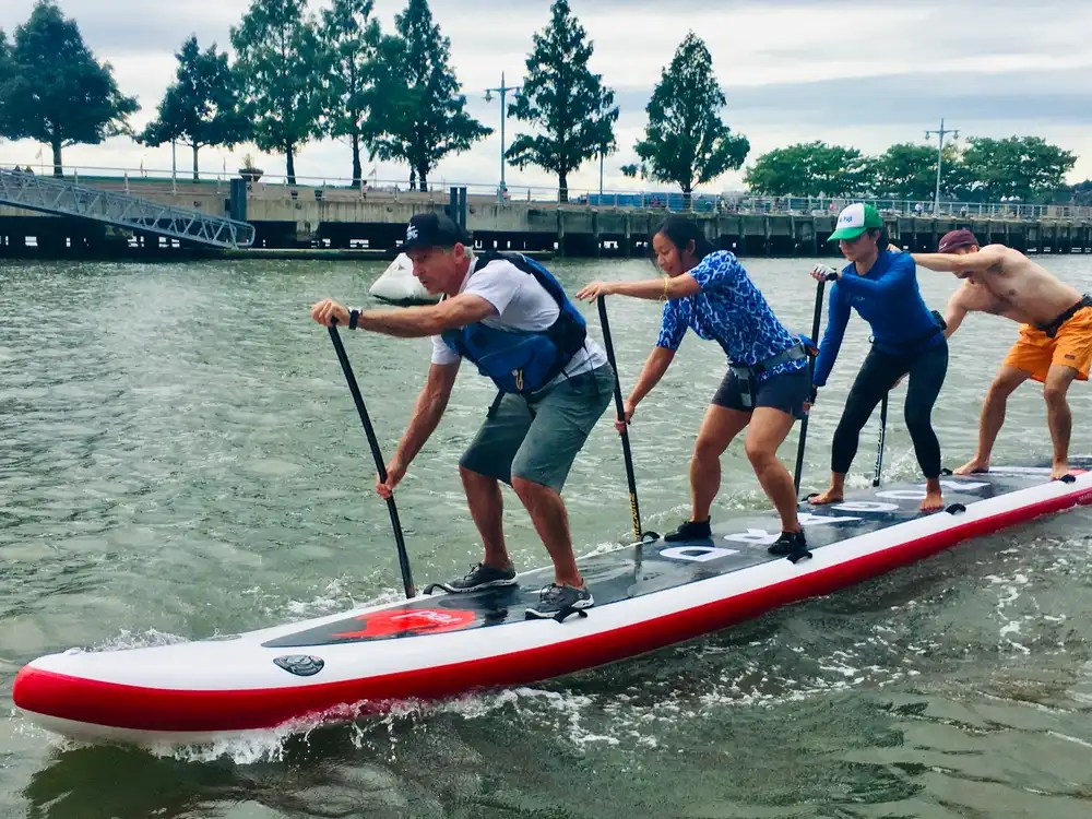 Four people paddle together on a large inflatable paddleboard on a river.