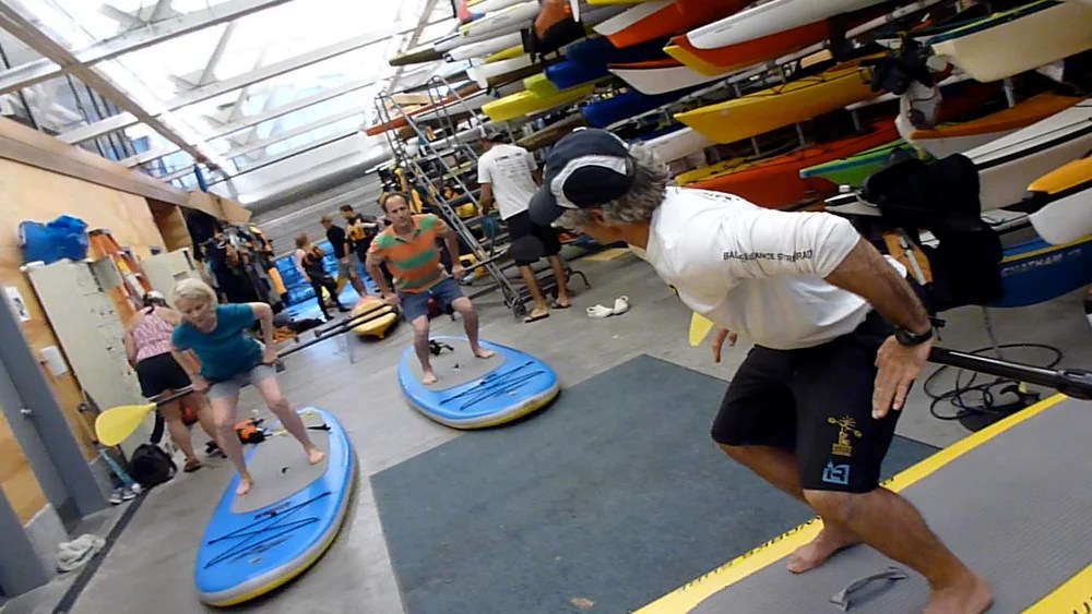 People standing on paddleboards indoors, practicing balancing.